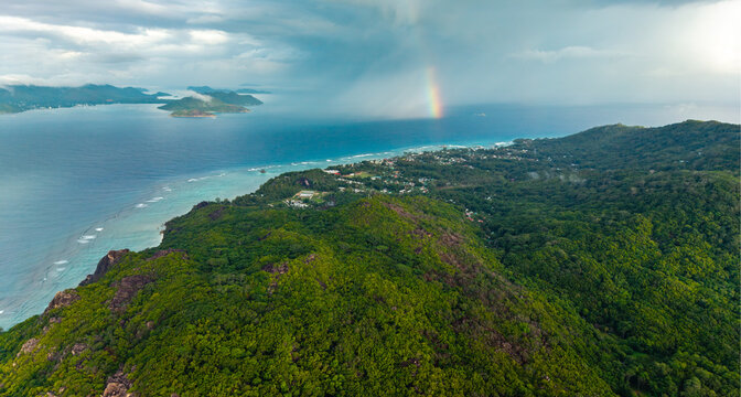 Aerial view of lush green mountains meet the turquoise sea under a vibrant rainbow's arc, Anse Reunion, La Digue, Seychelles.