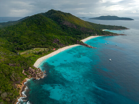 Aerial view of turquoise waters embracing the white sands of Anse Source d'Argent beach, nestled against a verdant coastline under a cloudy sky, Anse Reunion, La Digue, Seychelles.