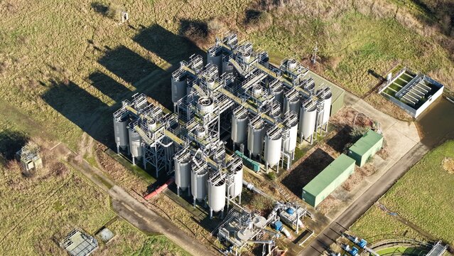 Aerial view of a sprawling sewage works complex with cylindrical tanks casting stark shadows on the surrounding fields, Luton, United Kingdom.