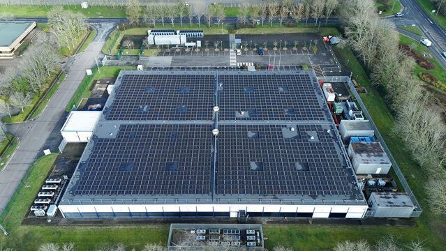 Aerial view of a sprawling data center roof adorned with a geometric array of solar panels, reflecting the sky amidst the urban landscape, Milton Keynes, United Kingdom.