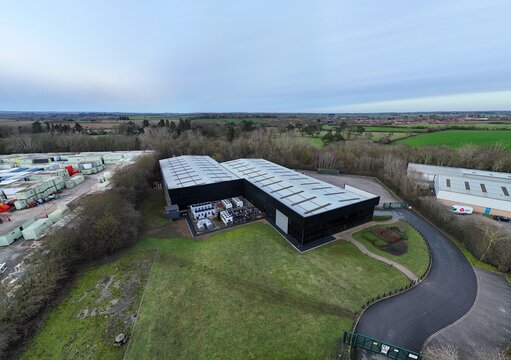 Aerial view of a modern, dark-colored building contrasting with the surrounding green landscape and distant bare trees, Node4 Data Centre, Northampton, United Kingdom.