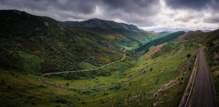 Aerial view of a winding road snakes through verdant hills under a dramatic sky, Aralla de Luna, Castilla y Leon, Spain.