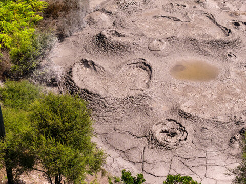 Aerial view of a geothermal landscape dotted with bubbling mud pools, contrasting with the lush greenery surrounding the textured earth, Rotorua, Bay of Plenty Region, New Zealand.