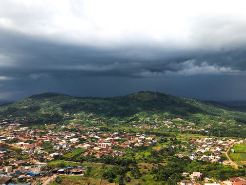 Aerial view of dark clouds looming over the lush green hills and small buildings, creating a striking contrast of light and shadow, Ilesa, Osun, Nigeria.