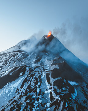 Aerial view of Mount Etna's peak ablaze against a serene sky, its snow-dusted slopes a stark contrast to the fiery eruption, Nicolosi, Sicily, Italy.