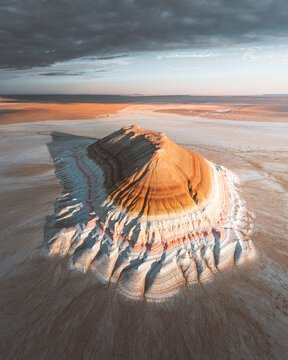 Aerial view of a majestic, layered mountain rising from the stark, sun-baked plains under a brooding sky, a symphony of earthy tones and dramatic shadows, Aktau, Mangystau Region, Kazakhstan.