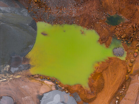 Aerial view of a vibrant, almost unnatural green pool contrasts sharply against the surrounding rust-colored earth, creating a surreal landscape, Gourlizon, Brittany, France.