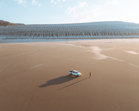 Aerial view of a lone figure standing near a white pickup truck on the vast, sun-baked sands beneath the towering, textured cliffs, Aktau, Mangystau Region, Kazakhstan.