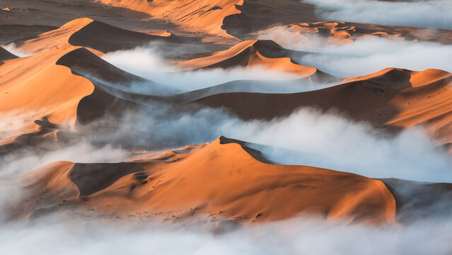 Aerial view of towering ochre dunes piercing through a sea of ethereal mist, creating a surreal landscape in the heart of the Namib Desert, Sossusvlei, Hardap Region, Namibia.