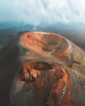 Aerial view of the volcanic landscape where the dark, rugged terrain meets the vibrant red crater, kissed by the soft touch of clouds, Nicolosi, Sicily, Italy.