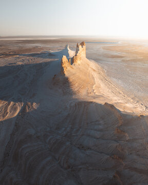 Aerial view of a dramatic rock formation rising from the desert floor, bathed in golden sunlight, creating stark shadows and highlighting the arid landscape, Aktau, Mangystau Region, Kazakhstan.