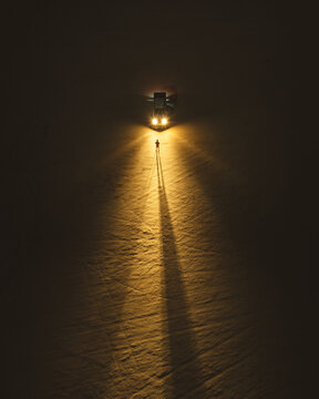 Aerial view of a lone figure casting a long shadow in the stark, golden glow of vehicle headlights, creating a mesmerizing contrast against the dark surroundings, Mangystau Region, Kazakhstan.