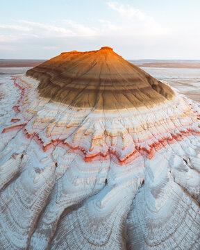Aerial view of the stark, striated beauty of a lone mountain, its layers of white, orange, and brown contrasting against the flat, pale landscape, Shetpe, Mangystau Region, Kazakhstan.