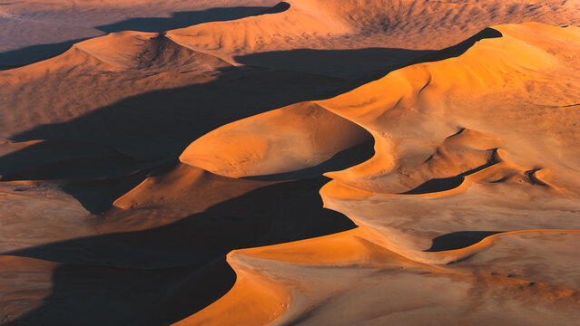 Aerial view of undulating dunes bask in the warm glow of the setting sun, casting long shadows across the sandy landscape, Sossusvlei, Hardap Region, Namibia.