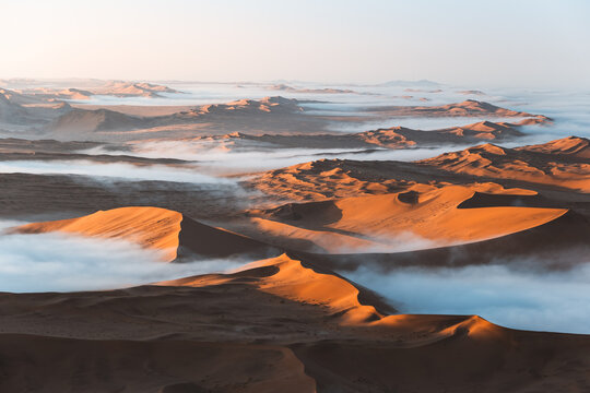 Aerial view of golden dunes piercing through a sea of ethereal fog, bathed in the soft glow of dawn, Sossusvlei, Hardap Region, Namibia.