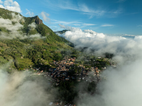 Aerial view of a quaint town nestled amid verdant mountains, partially veiled by ethereal clouds under a bright sky, Tamesis, Antioquia, Colombia.