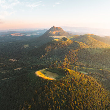 Aerial view of the verdant, rolling volcanic hills capped with golden sunlight contrasting against the deep green forests, Orcines, Auvergne-Rhone-Alpes, France.