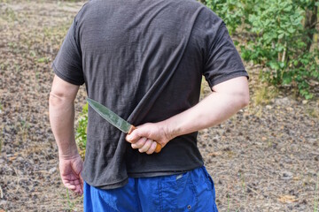 hand of a male criminal in black clothes and a gray knife behind his back on the street