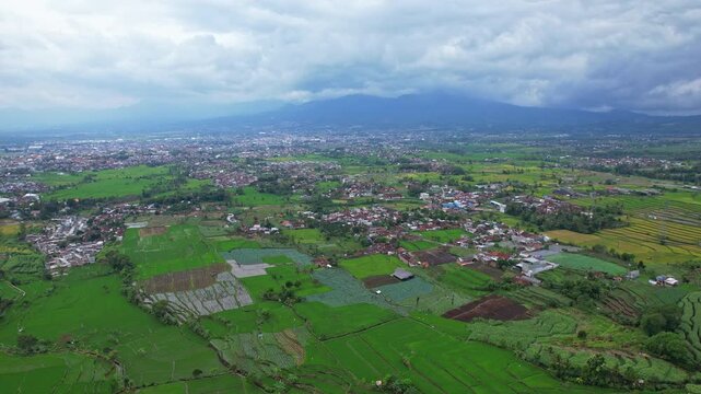 Aerial drone footage of a huge green agricultural valley, with many villages, surrounded by forests, rice fields, volcanic mountains, in cloudy atmosphere, in Garut area, Java island, Indonesia