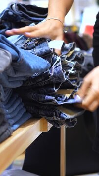 Female hands sorting through a stack of folded denim jeans on a wooden shelf, searching for the right size and style while shopping for new apparel in a retail fashion boutique