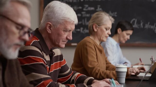 Group of older students sitting in row using laptops and collaborating in classroom with chalkboard in background