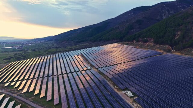 Rows of solar panels stretch across a field as the sun sets behind mountains. A small town is visible in the distance, highlighting renewable energy use in the area.