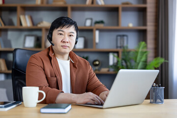 Asian man wearing a black headset sitting at a wooden desk with a laptop, typing and looking at the camera, working remotely from a modern home office with bookshelves © Liubomir