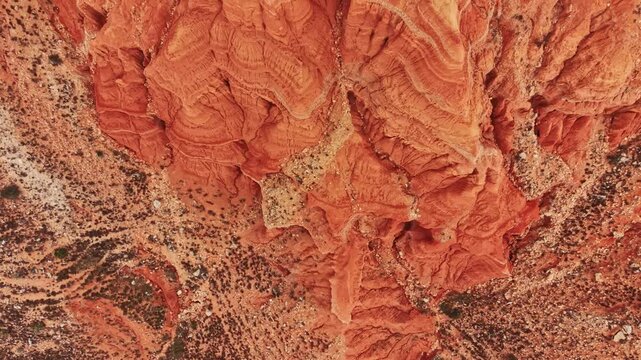 A wide view shows the canyon with layers of red rocks and interesting patterns. The ground is dry with some sparse vegetation. The sun brightens the scene.