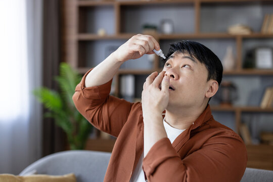 Asian man applying soothing eye drops, getting relief from dry eyes, allergies, or an eye condition, focusing on self-care and medical treatment at home