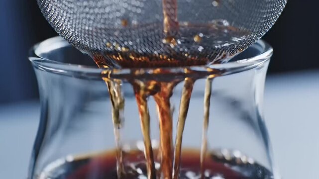 Pouring rich coffee liquid through a fine mesh strainer into a clear glass, capturing the brewing process