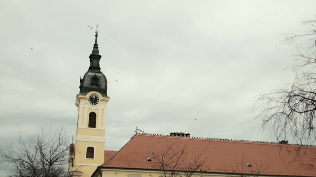 Historic church tower with clock and red tiled roof in Sombor, Serbia under cloudy winter sky. European architecture and cultural travel destination concept