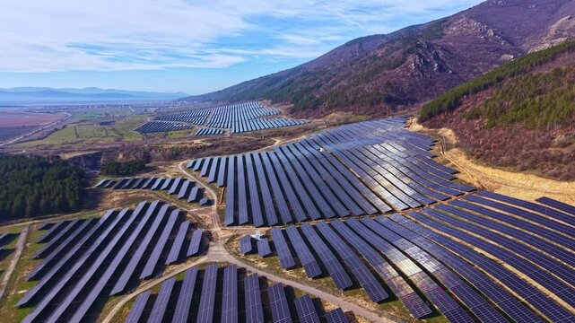 Rows of solar panels spread across a wide field near hills on a clear day. The bright sun reflects off the panels creating a shiny surface. The landscape shows green and brown land.