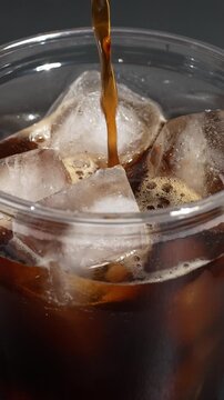 Iced coffee preparation process, coffee is poured into a plastic cup with ice on a dark background.