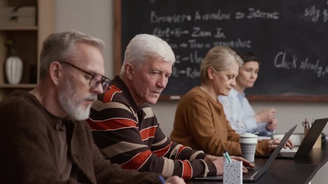 Older students sitting in row interacting and using laptops in classroom with chalkboard in background