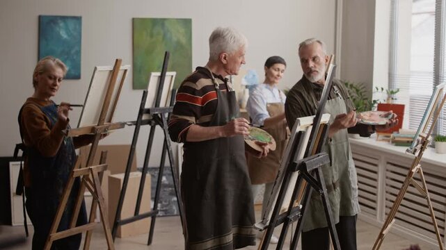 Group of senior people wearing aprons collaborating while painting on canvas in bright art studio