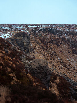 Rocky gritstone cliffs and moorland slopes with patchy snow under an overcast sky in the Peak District National Park, England. Atmospheric winter landscape with rugged textures and muted colours.