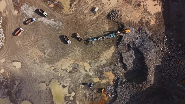 Heavy machinery and trucks operate at a granite quarry site. Workers extract stone from the ground. Trucks carry materials away from the site.