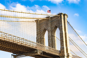 Fototapeta premium Brooklyn Bridge stone tower with American flag and steel cables against blue sky on a sunny day in New York City USA