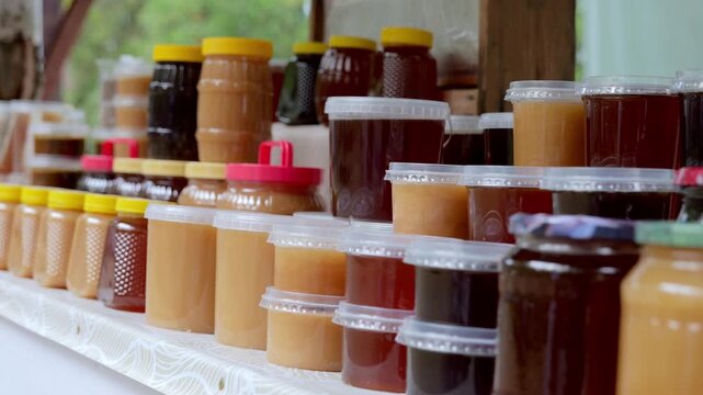 Farmhouse preserve setup, Beekeeper displays honey and jam jars on countrystyle table outdoors, Vivid scene of rustic table with assorted honey and jam jars arranged by beekeeper outdoors