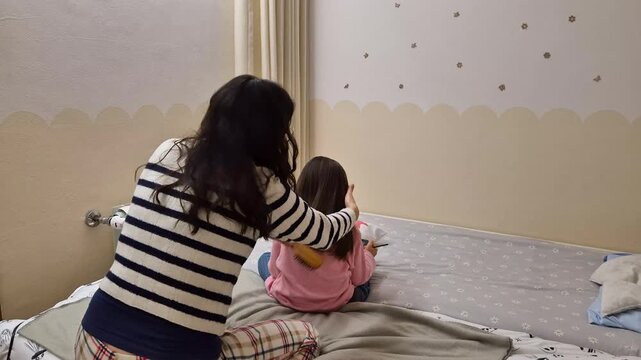 Mother brushing her daughter&rsquo;s long hair on a bed in a cozy children bedroom at the end of the day, showing real family daily hygiene routine, tenderness and personal care.