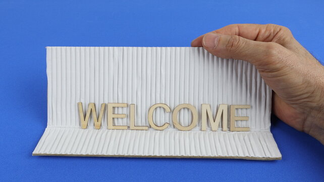 Un cartello di benvenuto in legno su cartone ondulato, tenuto dalla mano di un uomo su uno sfondo blu. Un concetto di benvenuto a casa.
