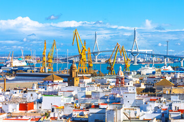 Cityscape showcases historic white rooftops of Cadiz, Spain, set against a modern industrial backdrop. Massive yellow shipyard cranes and the towering Bridge dominate the horizon over the bay © Leilani