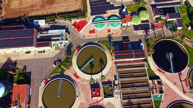 The picture shows a water treatment site with circular tanks and solar panels. There are pathways, plants, and clear areas. It is bright and sunny.