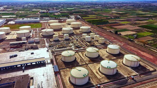 A large oil storage facility shows many big tanks organized in rows. The site is near fields and has roads with trucks delivering supplies. The day is bright and clear.