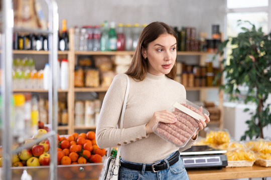 Young woman in store examines raw bbg sausage, chooses raw meat for dinner. Buyer examines information on label, examines condition of food.