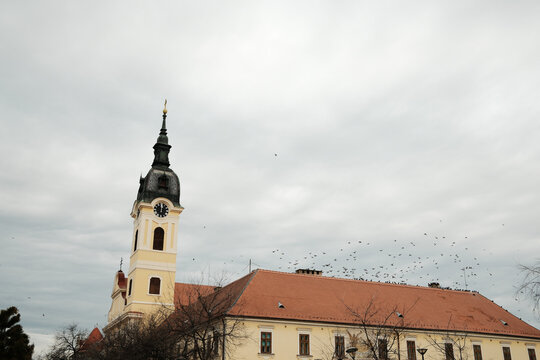 Church tower with clock and red roof under cloudy sky in Sombor, Serbia. European landmark with birds in flight and heritage concept.