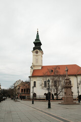 Obraz premium Church with clock tower and stone cross monument on open square in Sombor, Serbia. European religious heritage and city landmark concept.