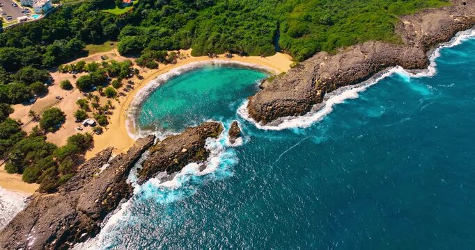 Turquoise water sparkling in the sun reaches the sandy beach through the gap in the rocks. Lush greenery surrounds Manati Beach in Puerto Rico. Aerial view.