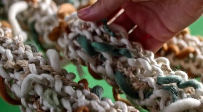 ​A close-up shot of an intricate handmade macrame or woven textile featuring thick, intertwined yarn in natural earthy tones of beige, brown, and teal against a green background.