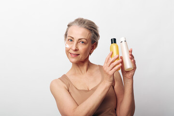 Fototapeta premium Mature woman with natural skin holds two cosmetic bottles, smiling gently. Skincare, beauty, and wellness concept focused on aging care and natural facial treatment on white background.